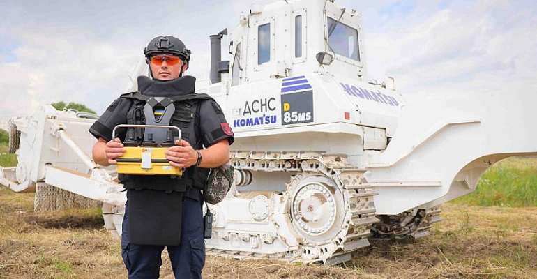 A Ukrainian deminer in full protective gear and a helmet stands in a field holding a detection device, with a large white armored Komatsu bulldozer in the background. The scene depicts landmine clearance operations in Ukraine, supported by the UNDP. 